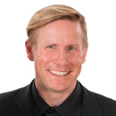 A professional headshot of a smiling man in a black suit.