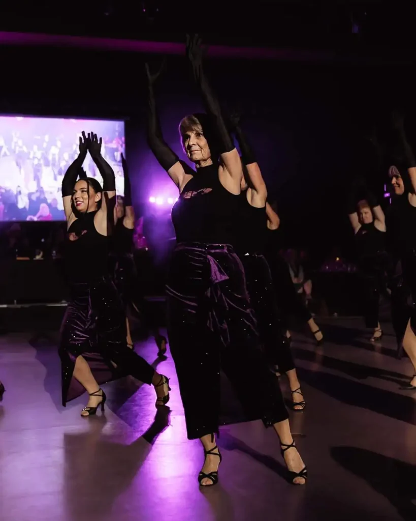 A group of women in black and purple outfits performing a synchronized dance under purple lighting.