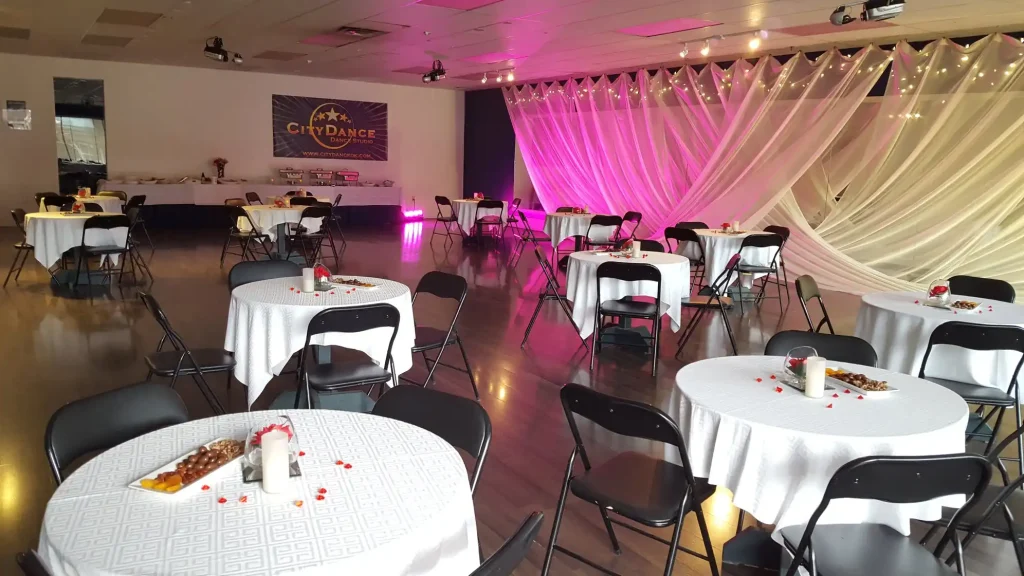 CityDance Studio event venue with round tables covered in white cloths, decorated with candles and snacks, under pink and purple lighting, 2025.