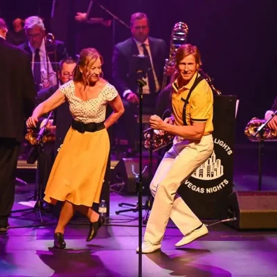 A lively couple dancing on stage with a jazz band in the background under purple lighting.